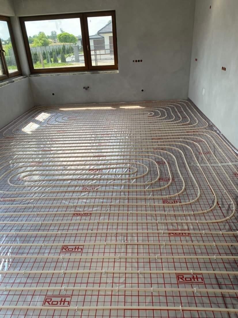Rozłożone rury ogrzewania podłogowego marki Roth na folii izolacyjnej z czerwoną siatką w surowym wnętrzu z oknem.