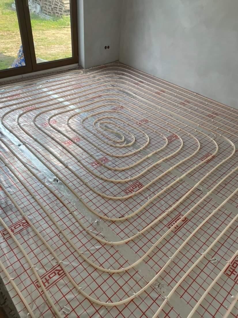 Rozłożona instalacja ogrzewania podłogowego Roth na folii z czerwoną siatką w jasnym pomieszczeniu z oknem i surowymi ścianami.