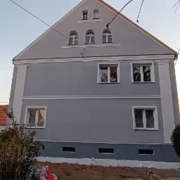 Szary budynek z czerwonym dachem po renowacji elewacji, widoczne detale architektoniczne i świeżo pomalowana fasada z elementami dekoracyjnymi.