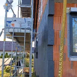Ściana budynku z cegły ceramicznej w trakcie docieplania szarymi płytami izolacyjnymi, widoczne rusztowanie i żółty przewód wentylacyjny.