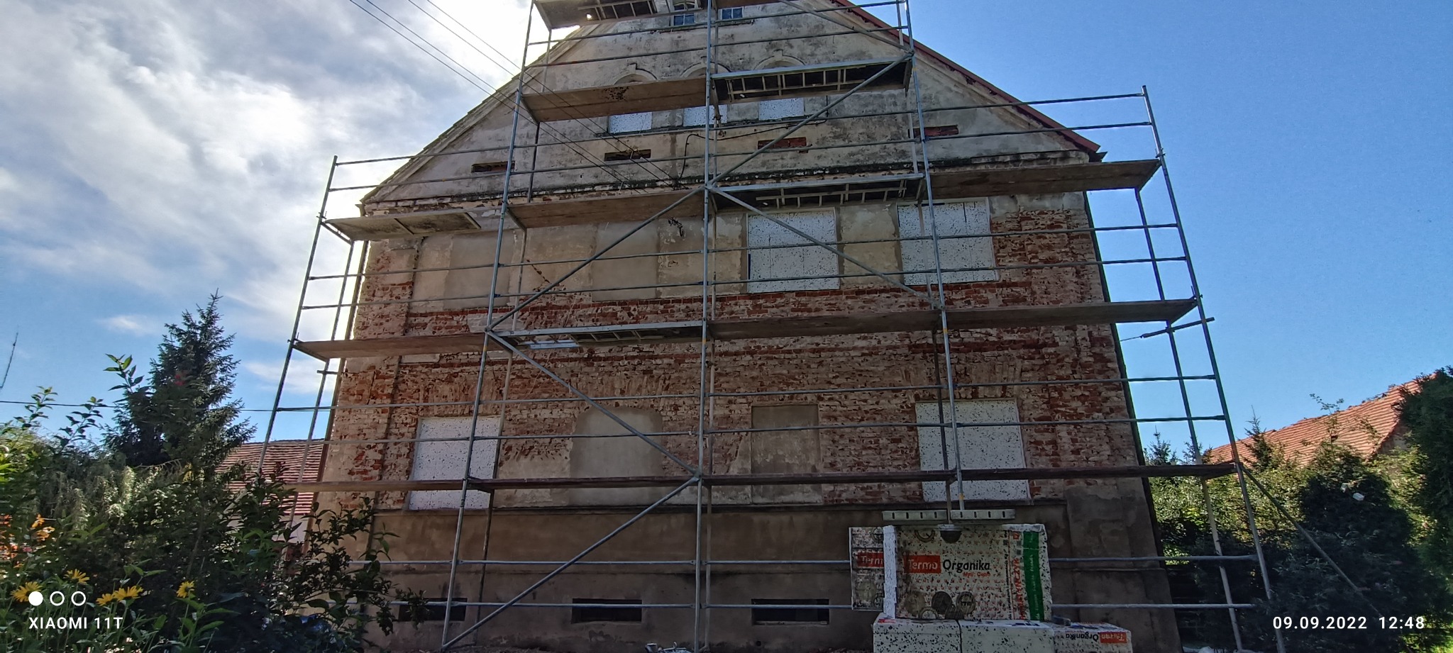 Stara, ceglana kamienica w trakcie docieplania, z widocznymi rusztowaniami i częściowo zamontowanymi płytami termoizolacyjnymi na elewacji, oraz stosem materiałów izolacyjnych u podstawy ściany.