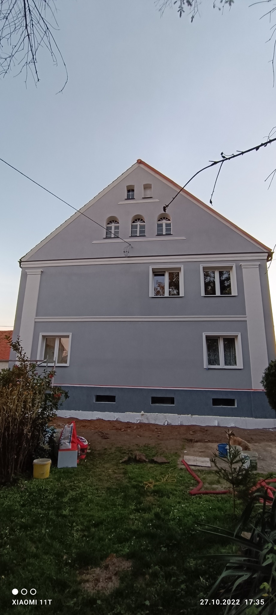 Szary budynek z czerwonym dachem po renowacji elewacji, widoczne detale architektoniczne i świeżo pomalowana fasada z elementami dekoracyjnymi.