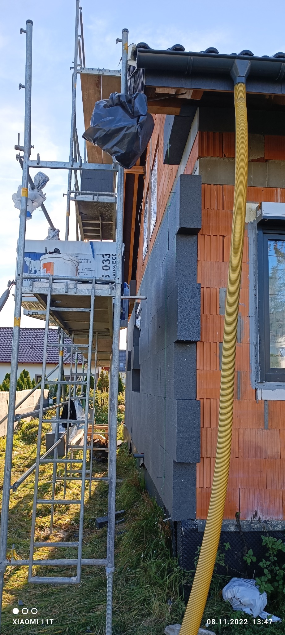 Ściana budynku z cegły ceramicznej w trakcie docieplania szarymi płytami izolacyjnymi, widoczne rusztowanie i żółty przewód wentylacyjny.