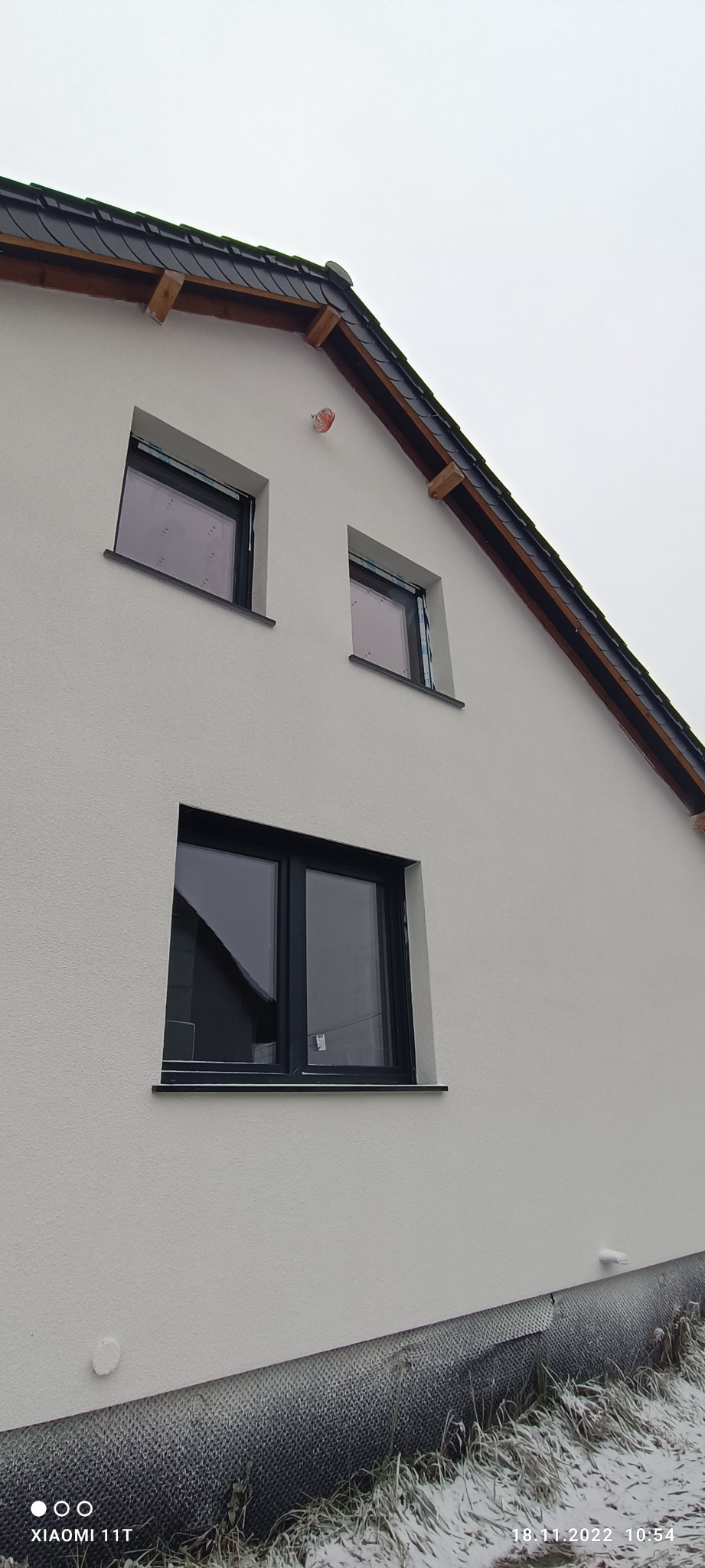 Fragment ocieplonej elewacji budynku z trzema czarnymi oknami, widoczne zabezpieczenia na oknach, częściowo ośnieżona trawa u podstawy ściany, pochmurne niebo.