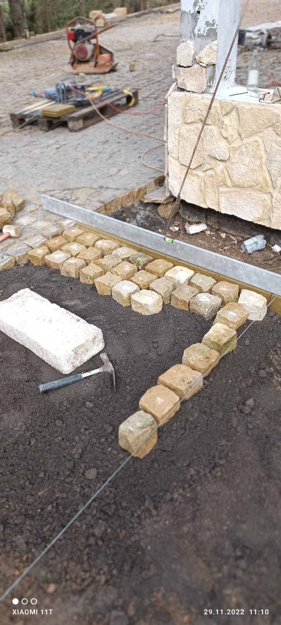 Układanie kostki brukowej z granitu przy budynku z kamienną elewacją, widoczny poziomica, sznurek traserski, młotek i ubijarka w tle.