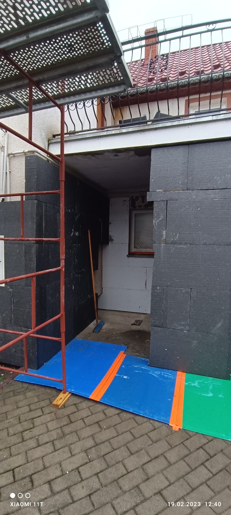 Montaż czarnych płyt izolacyjnych na elewacji budynku, widoczne rusztowanie i zabezpieczenia podłoża folią w kolorach niebieskim i zielonym.