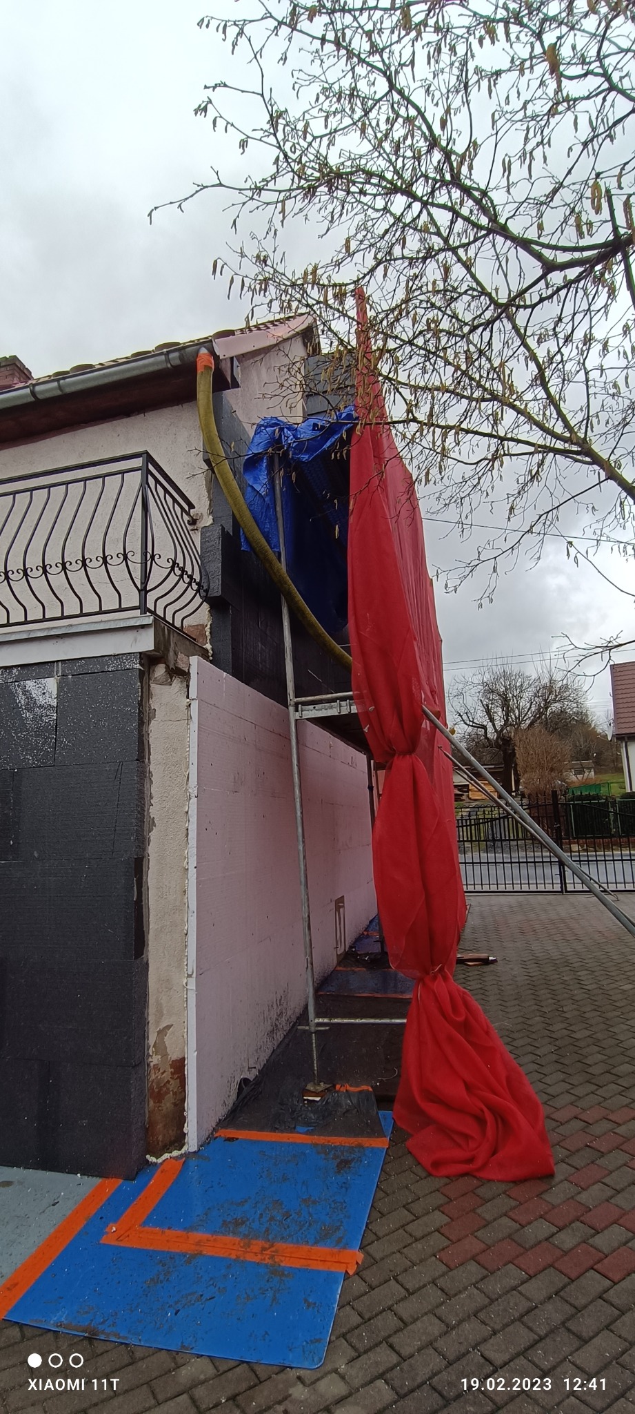 Ocieplanie elewacji domu jednorodzinnego w trakcie realizacji, widoczne rusztowanie z czerwoną plandeką ochronną, fragment ocieplonej ściany w kolorze grafitowym oraz fragment ściany z różową...