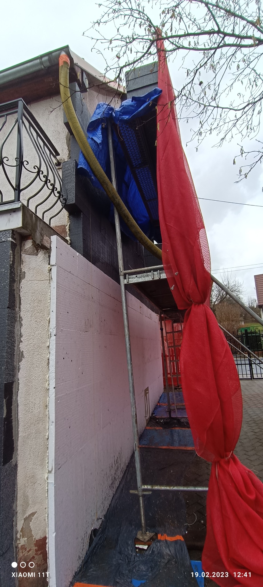 Ściana budynku w trakcie prac, z widocznym ociepleniem styropianem, rusztowaniem zabezpieczonym czerwoną siatką i niebieską plandeką, oraz żółtym wężem wentylacyjnym.
