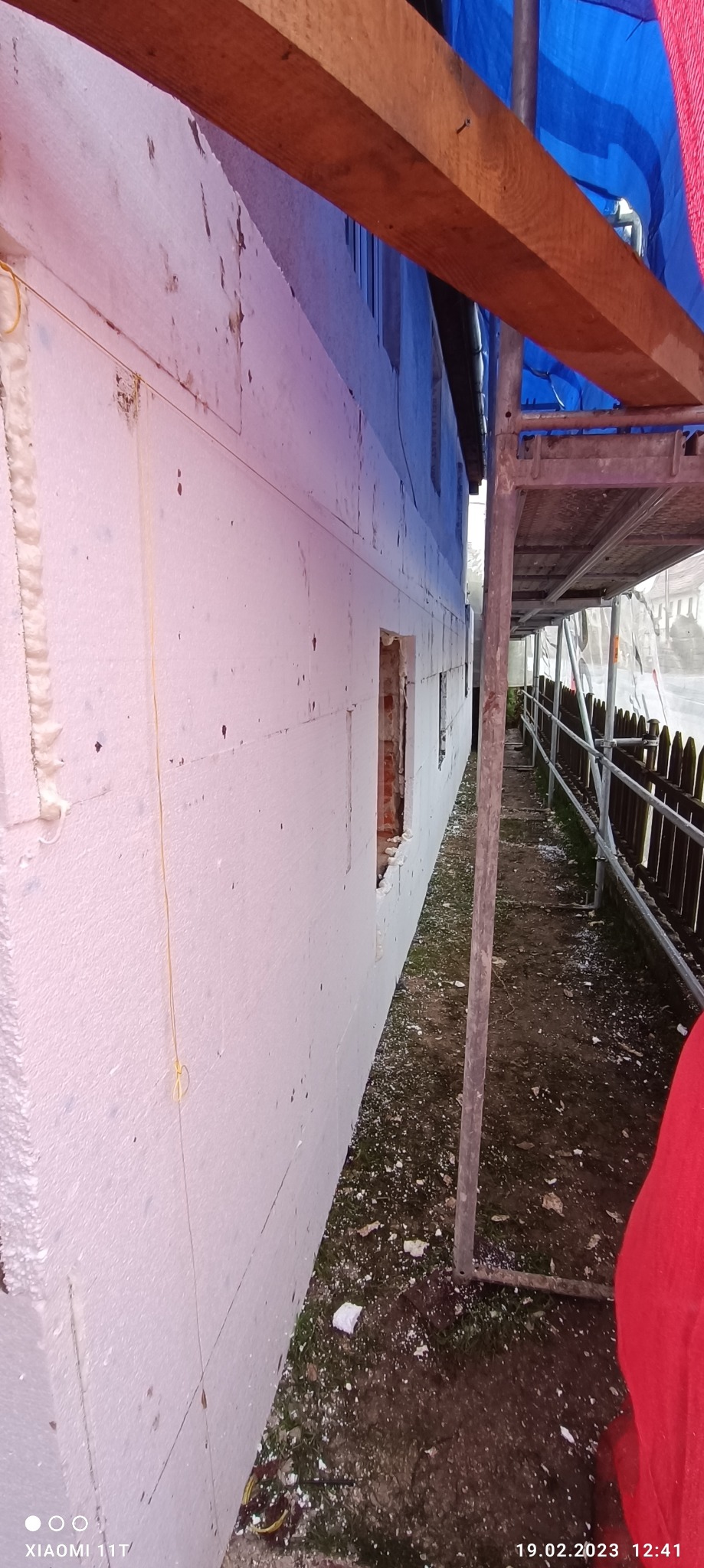 Ściana budynku w trakcie nakładania białego styropianu jako izolacji termicznej, widoczne rusztowanie i fragment okna bez ramy, z pozostałościami cegieł.