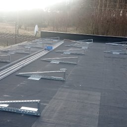 Avoltaic Kamil Kopaniecki - Montaż konstrukcji wsporczej pod panele fotowoltaiczne na płaskim dachu pokrytym papą, widoczne szyny montażowe, uchwyty trójkątne, butla gazowa i pracownik w trakcie mocowania elementów.