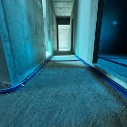 Avoltaic Kamil Kopaniecki - Wnętrze w trakcie prac: korytarz z surowymi ścianami, podłogą i niebieskimi rurami instalacyjnymi ułożonymi wzdłuż ścian. Widok w perspektywie, z oknem w tle.