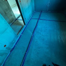 Avoltaic Kamil Kopaniecki - Niebieskie rury instalacji elektrycznej ułożone w surowym wnętrzu betonowego budynku przed wylaniem posadzki, widoczne uchwyty mocujące rury do podłoża.