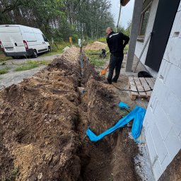 Avoltaic Kamil Kopaniecki - Wykopany rów pod budynkiem z widocznymi niebieskimi rurami, pracownik z łopatą i zaparkowany biały bus w tle. Prace ziemne przy budynku.