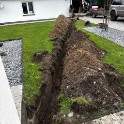 Avoltaic Kamil Kopaniecki - Wykop w trawniku pod kabel, widoczny czarny przewód w ziemi. W tle budynek z numerem 24, minikoparka i osoby nadzorujące prace.