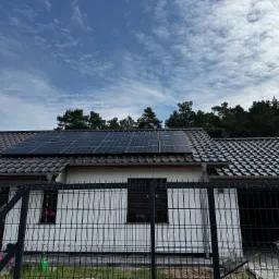 Dom jednorodzinny z panelami fotowoltaicznymi na dachu, widok zza ogrodzenia panelowego. Błękitne niebo z chmurami w tle. Ujęcie z poziomu gruntu.