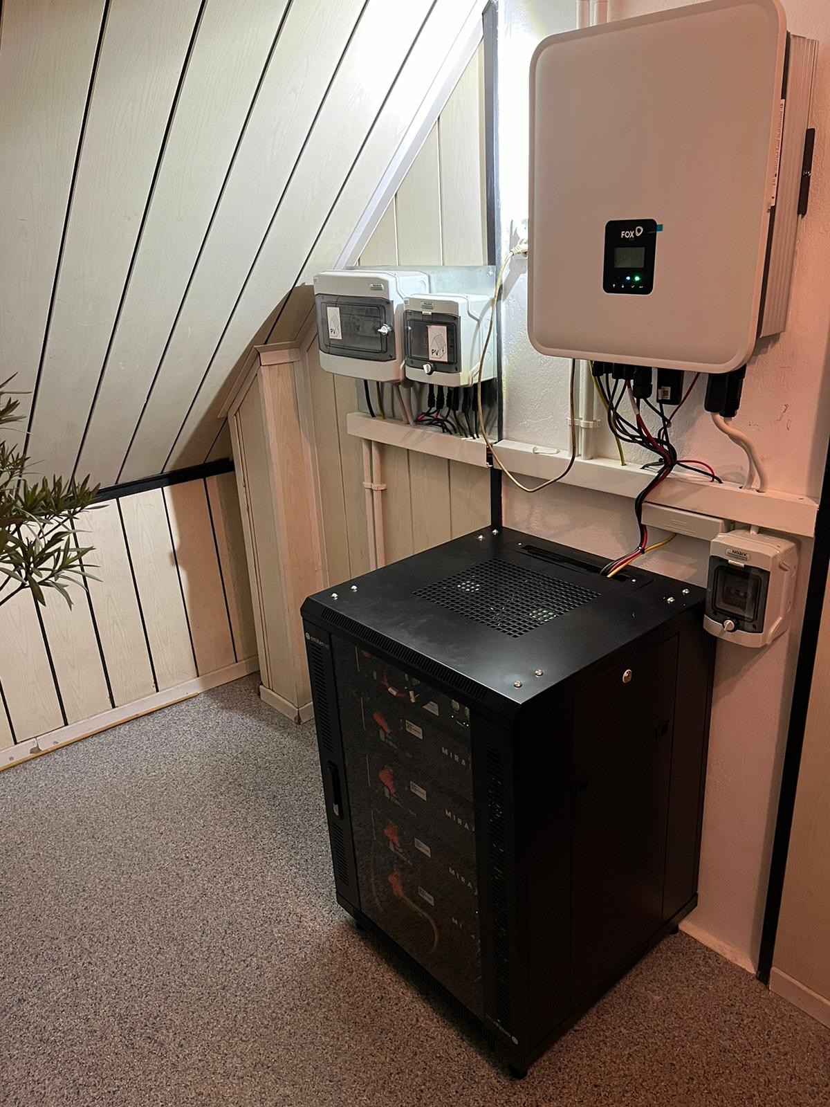 Wnętrze pomieszczenia z zamontowanym inwerterem fotowoltaicznym Fox ESS, rozdzielnicami elektrycznymi i czarną szafą rack z okablowaniem na ścianie z boazerią.