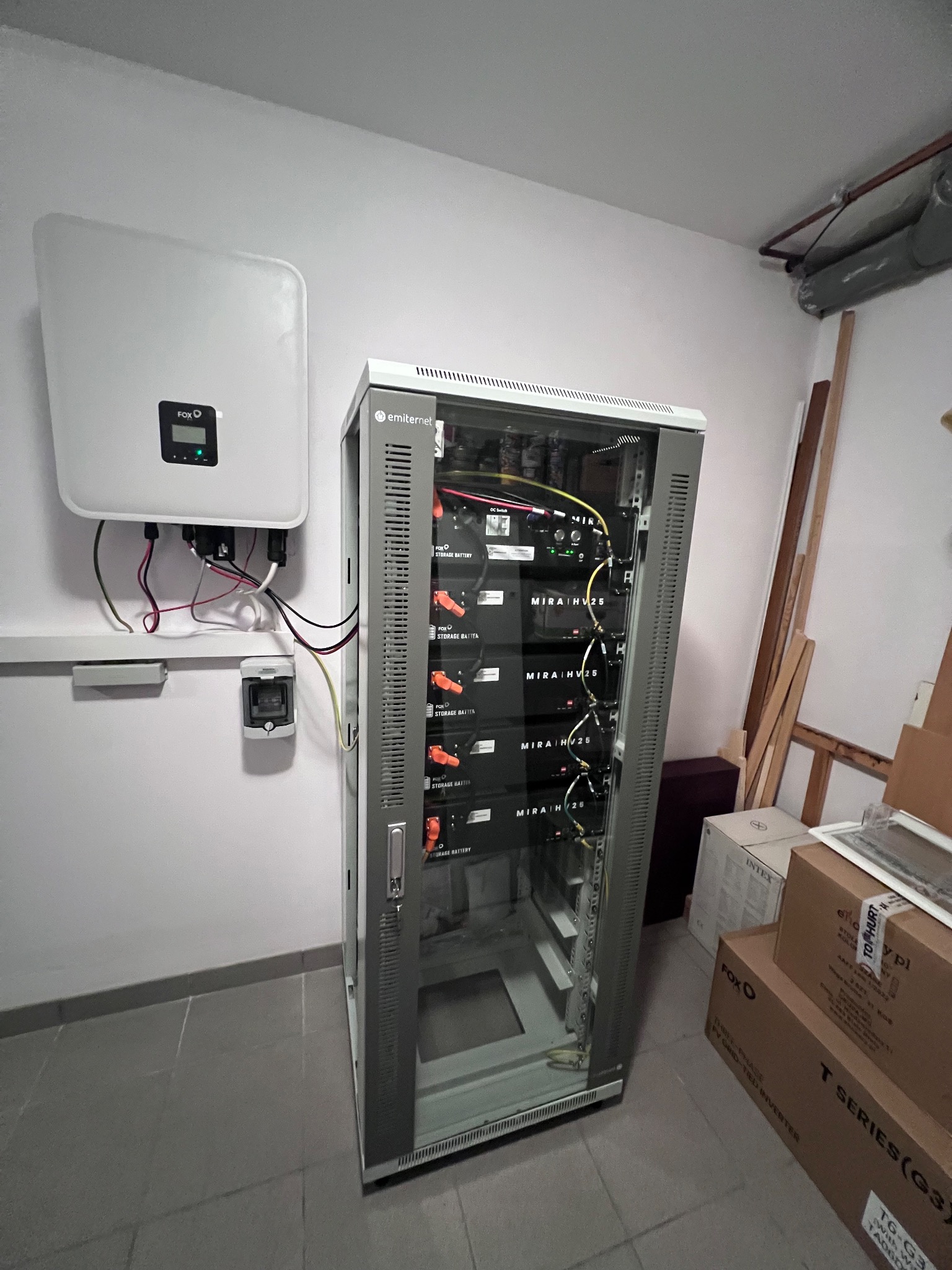 Instalacja fotowoltaiczna w pomieszczeniu: inwerter na ścianie, obok szafa rack z bateriami magazynującymi energię, widoczne okablowanie i skrzynka elektryczna pod inwerterem.