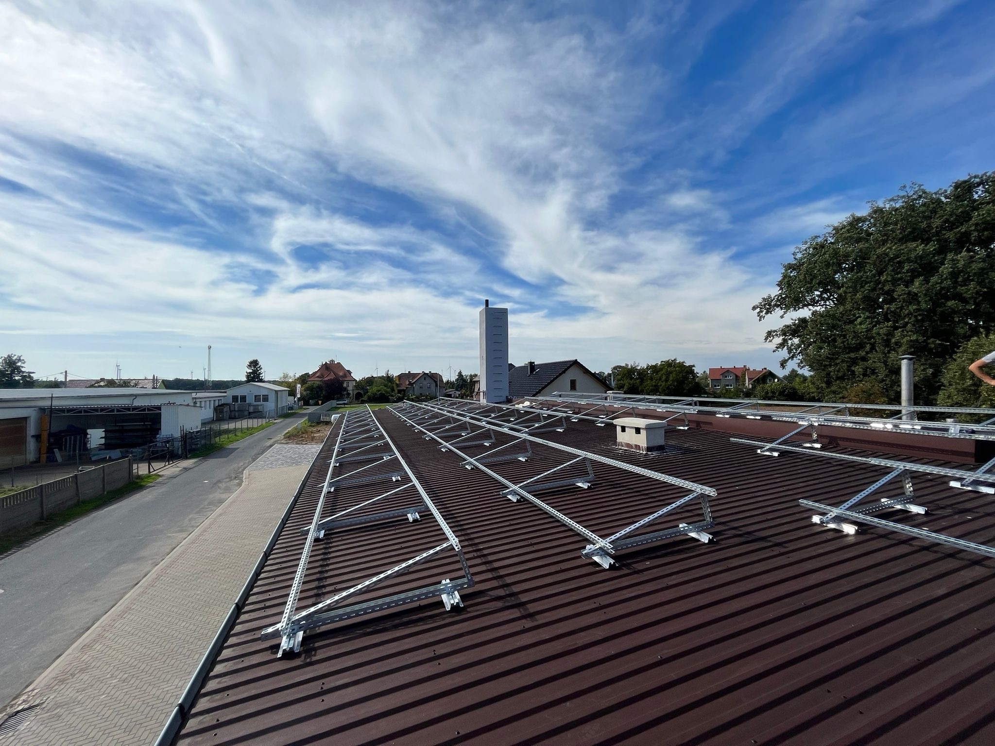 Metalowa konstrukcja wsporcza pod panele fotowoltaiczne zamontowana na brązowym dachu z blachy trapezowej, widok z góry, błękitne niebo z chmurami w tle.