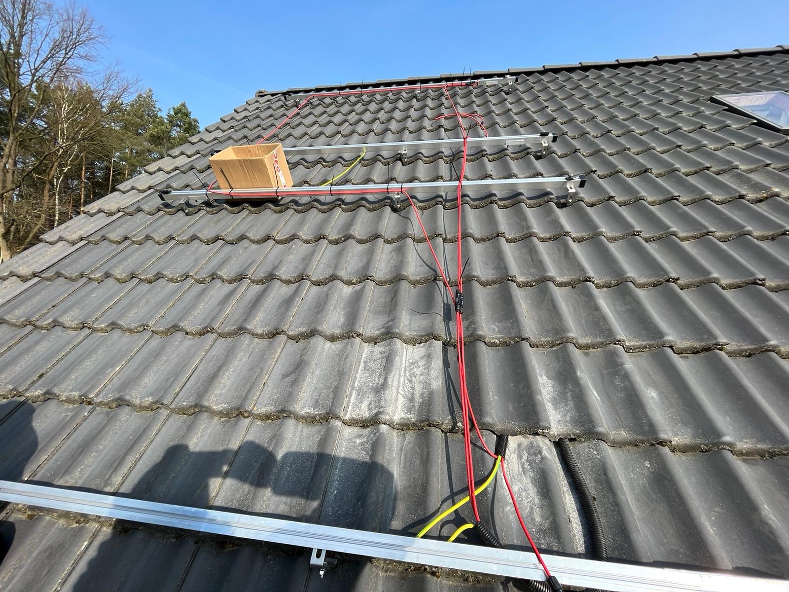 Montaż paneli słonecznych na dachu z dachówki ceramicznej; widoczne szyny montażowe, przewody elektryczne w kolorach czerwonym i żółtym, oraz karton na dachu.