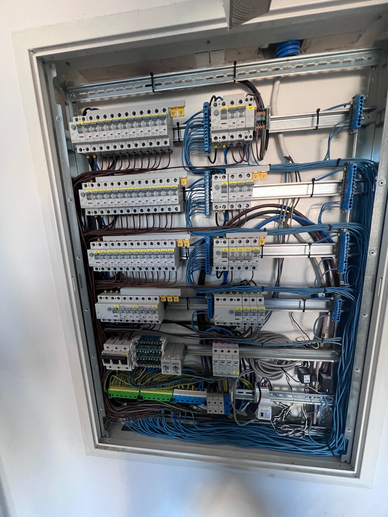 Rozdzielnia elektryczna w metalowej obudowie, widoczne liczne bezpieczniki, przewody w kolorze niebieskim i brązowym, listwy zaciskowe, zamocowane na szynach DIN w białym wnętrzu szafki.