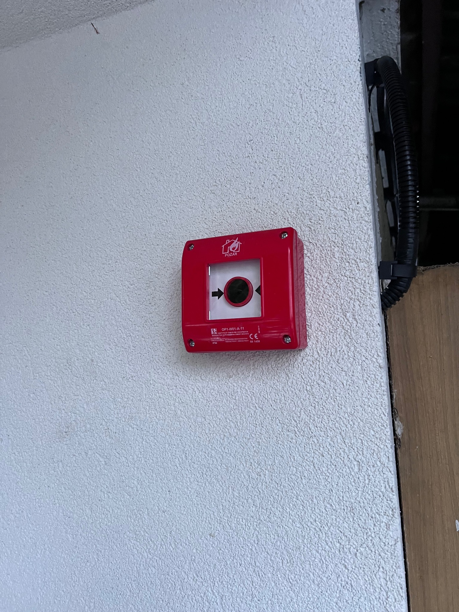 Czerwony, kwadratowy przycisk alarmowy przeciwpożarowy z czarną strzałką i symbolem płomienia, zamontowany na teksturowanej, białej ścianie w pobliżu rury osłonowej kabli elektrycznych.