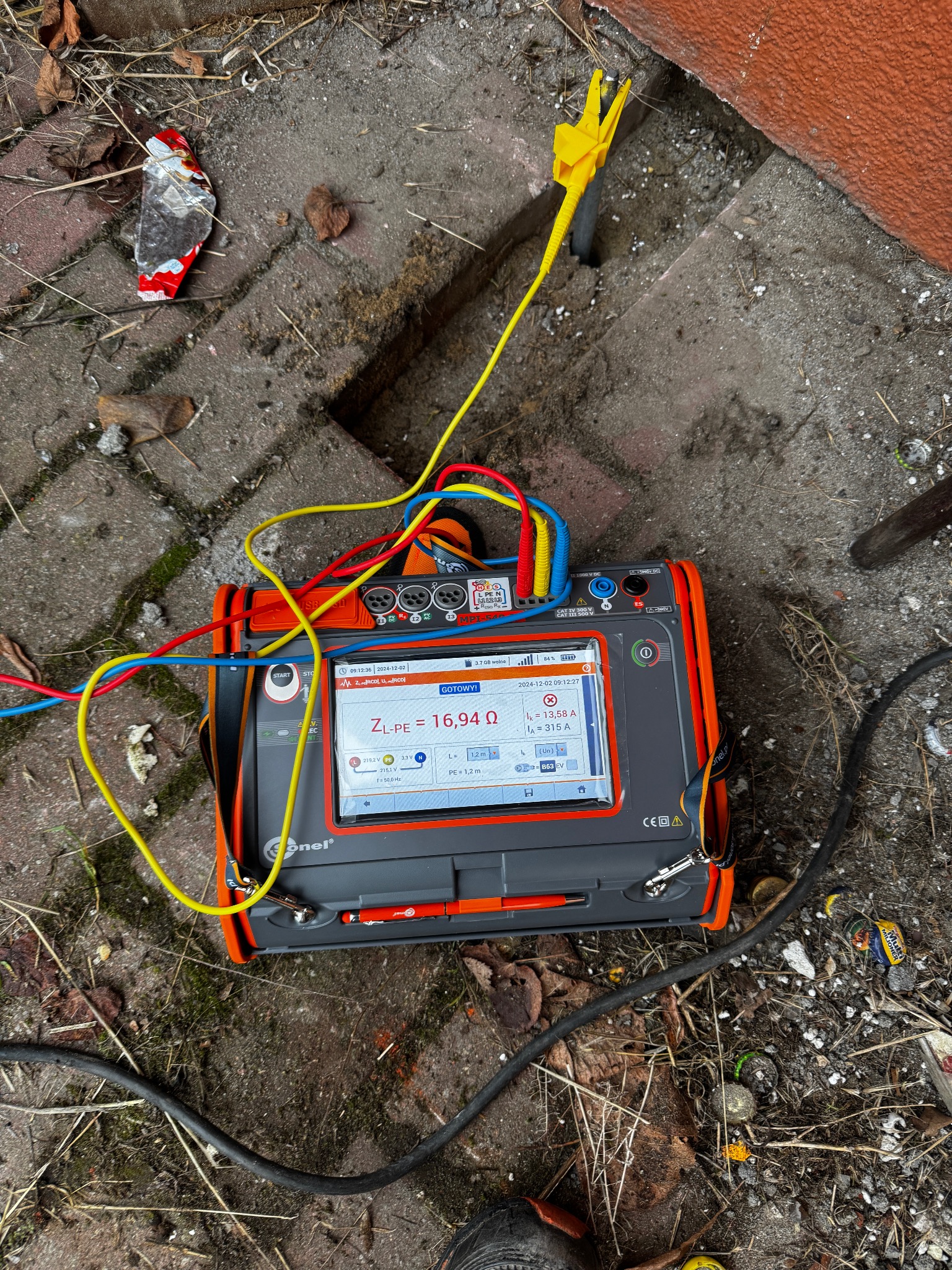 Miernik elektryczny Sonel MPI-54 z podłączonymi przewodami pomiarowymi, wyświetlający wynik pomiaru impedancji pętli zwarcia ZL-PE na brukowanym podłożu z widocznymi śladami ziemi i drobnych śmieci.