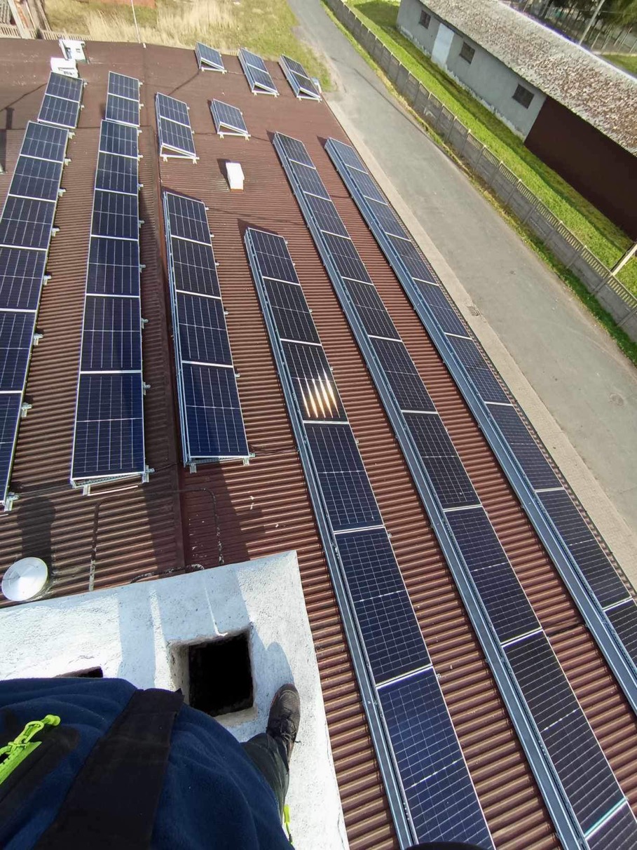 Widok z góry na instalację paneli słonecznych na dachu z blachy trapezowej, z perspektywy osoby stojącej na kominie, widoczne fragmenty ubrania roboczego i buta.