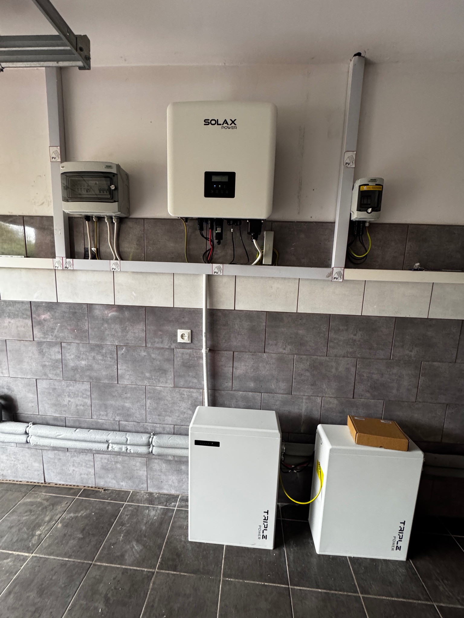 Kompletna instalacja fotowoltaiczna w garażu: inwerter Solax Power, dwa akumulatory Triple Power, rozdzielnica i zabezpieczenia. Szare płytki na ścianie i podłodze.