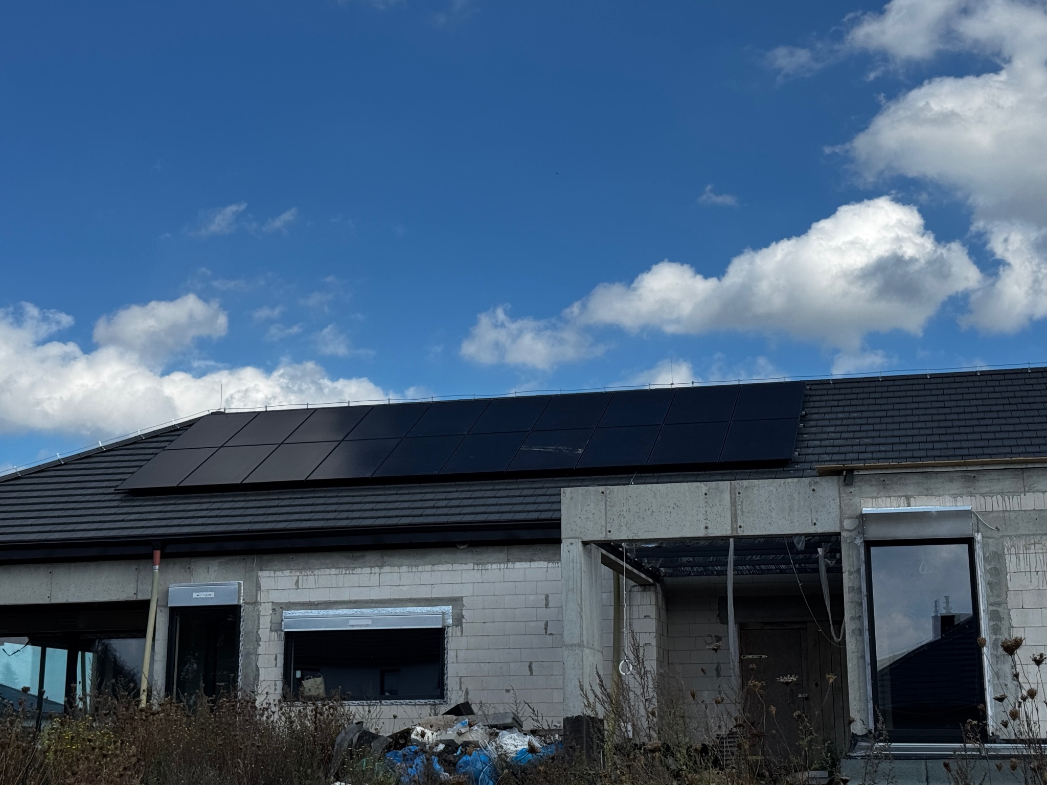 Dom w trakcie budowy z zamontowanymi panelami fotowoltaicznymi na dachu. Błękitne niebo z chmurami w tle.