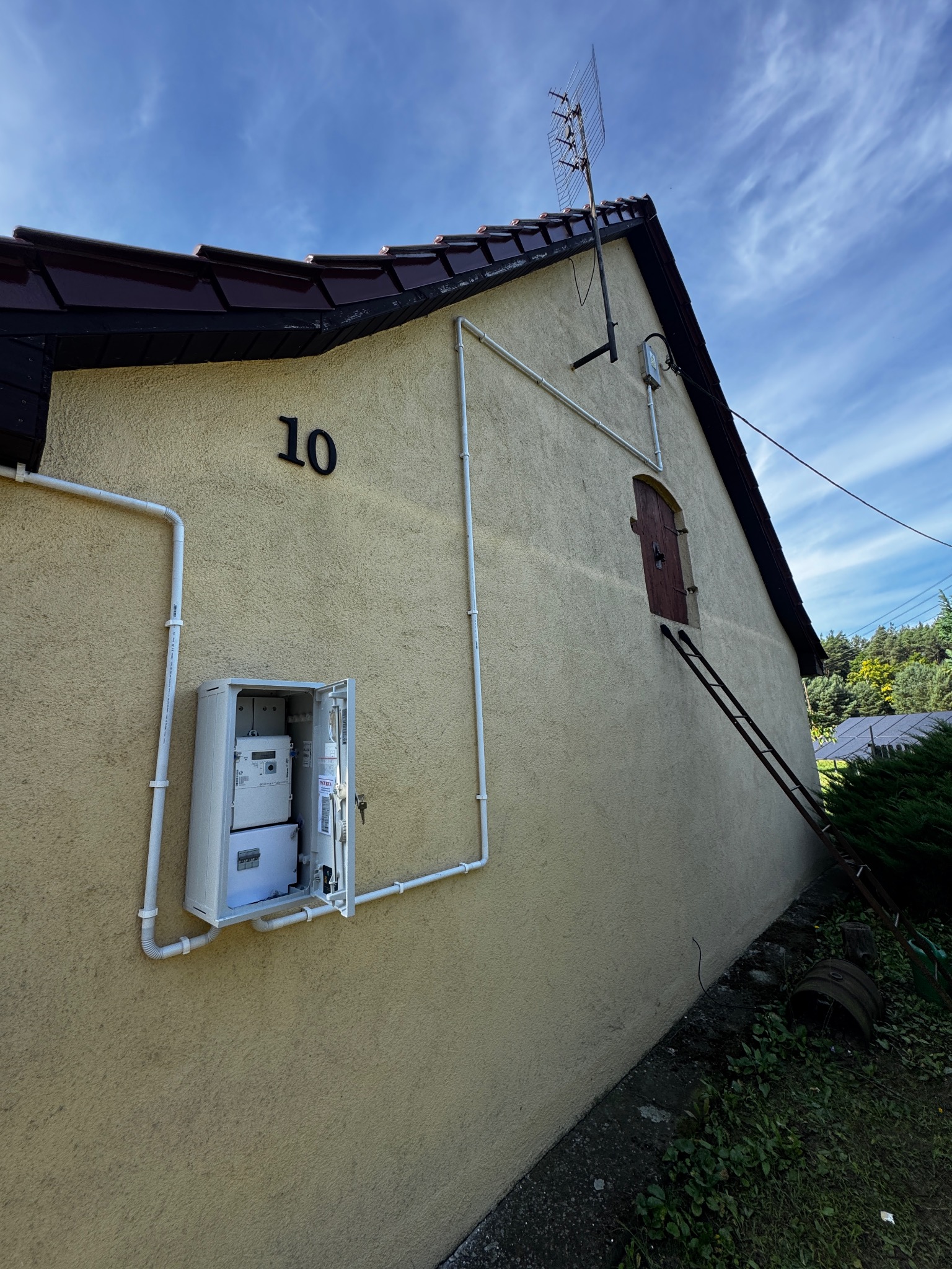 Elewacja budynku z numerem 10, skrzynką licznikową i instalacją elektryczną. Antena na dachu i drabina oparta o ścianę. Widok pod kątem.