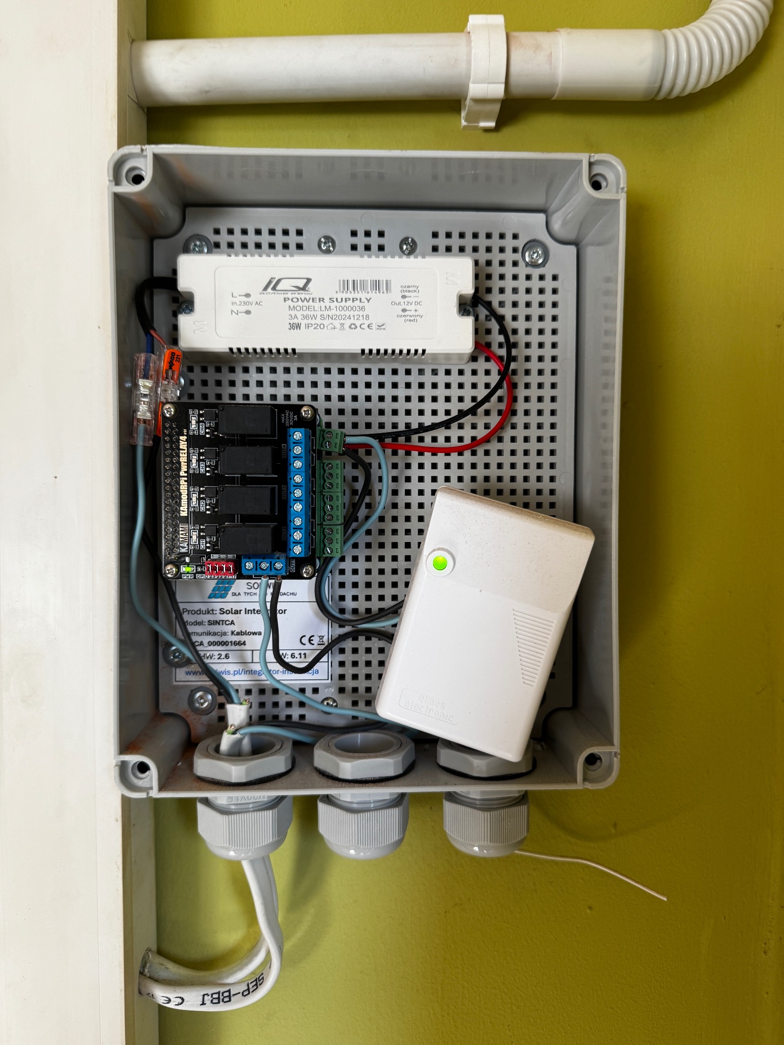 Szara skrzynka elektryczna z modułami, w tym zasilaczem IQ Power Supply i płytką Kamami, zamontowana na ścianie. Widoczne kable i oznaczenia producentów.