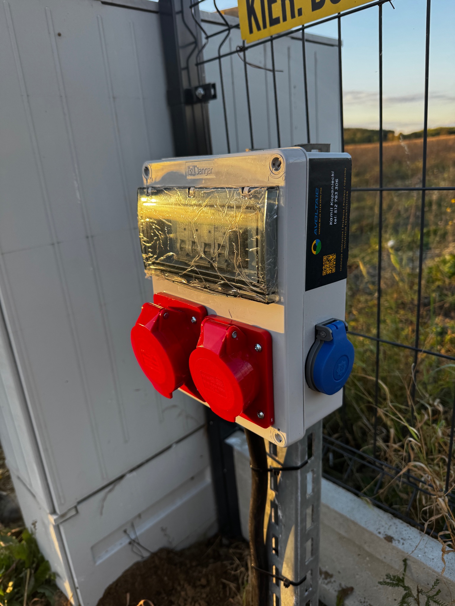 Szara skrzynka z dwoma czerwonymi gniazdami i jednym niebieskim, zamontowana na metalowym słupku przy ogrodzeniu. Widoczna tabliczka z danymi firmy Avoltaic.