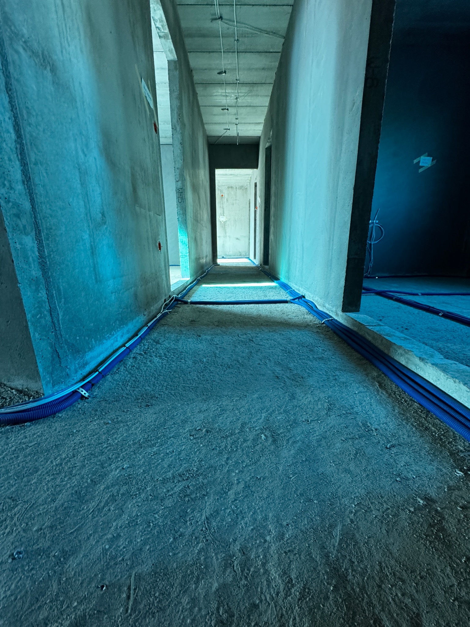 Wnętrze w trakcie prac: korytarz z surowymi ścianami, podłogą i niebieskimi rurami instalacyjnymi ułożonymi wzdłuż ścian. Widok w perspektywie, z oknem w tle.