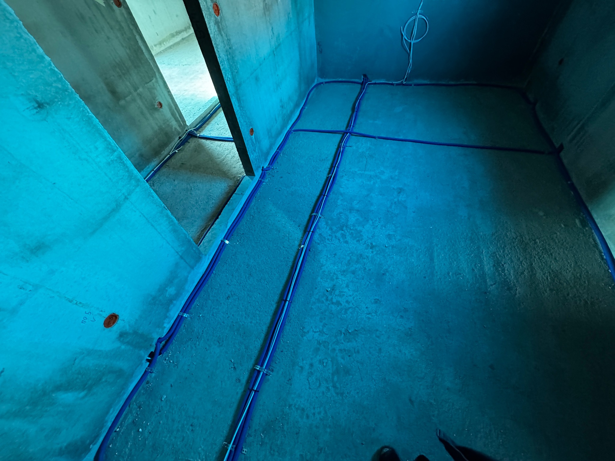 Niebieskie rury instalacji elektrycznej ułożone w surowym wnętrzu betonowego budynku przed wylaniem posadzki, widoczne uchwyty mocujące rury do podłoża.