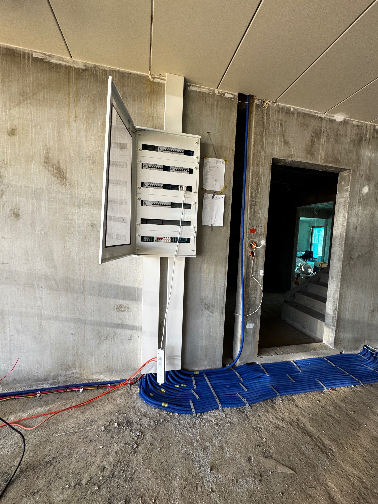 Otwarta skrzynka rozdzielcza z widocznymi zabezpieczeniami, kable w niebieskich peszlach ułożone na podłodze w surowym pomieszczeniu w trakcie instalacji elektrycznej.