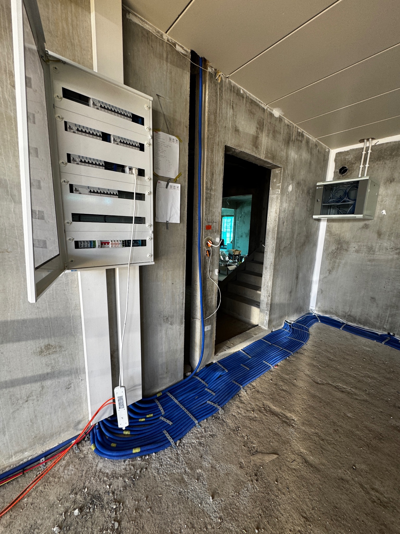 Rozdzielnia elektryczna z otwartymi drzwiczkami na surowej ścianie. Widoczne niebieskie rury instalacyjne na podłodze w szarym pomieszczeniu w trakcie wykańczania.
