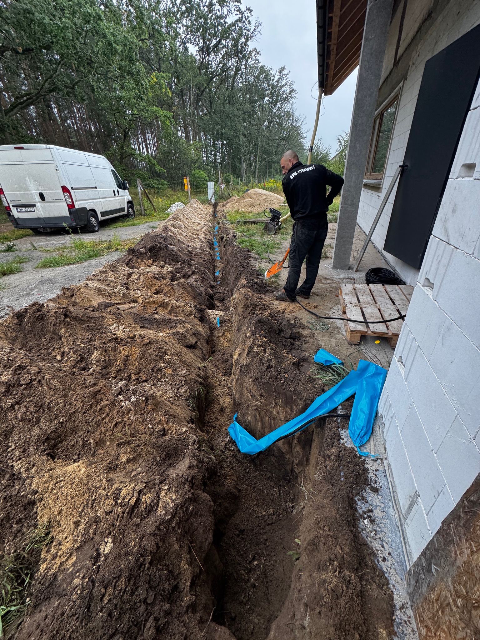 Wykopany rów pod budynkiem z widocznymi niebieskimi rurami, pracownik z łopatą i zaparkowany biały bus w tle. Prace ziemne przy budynku.
