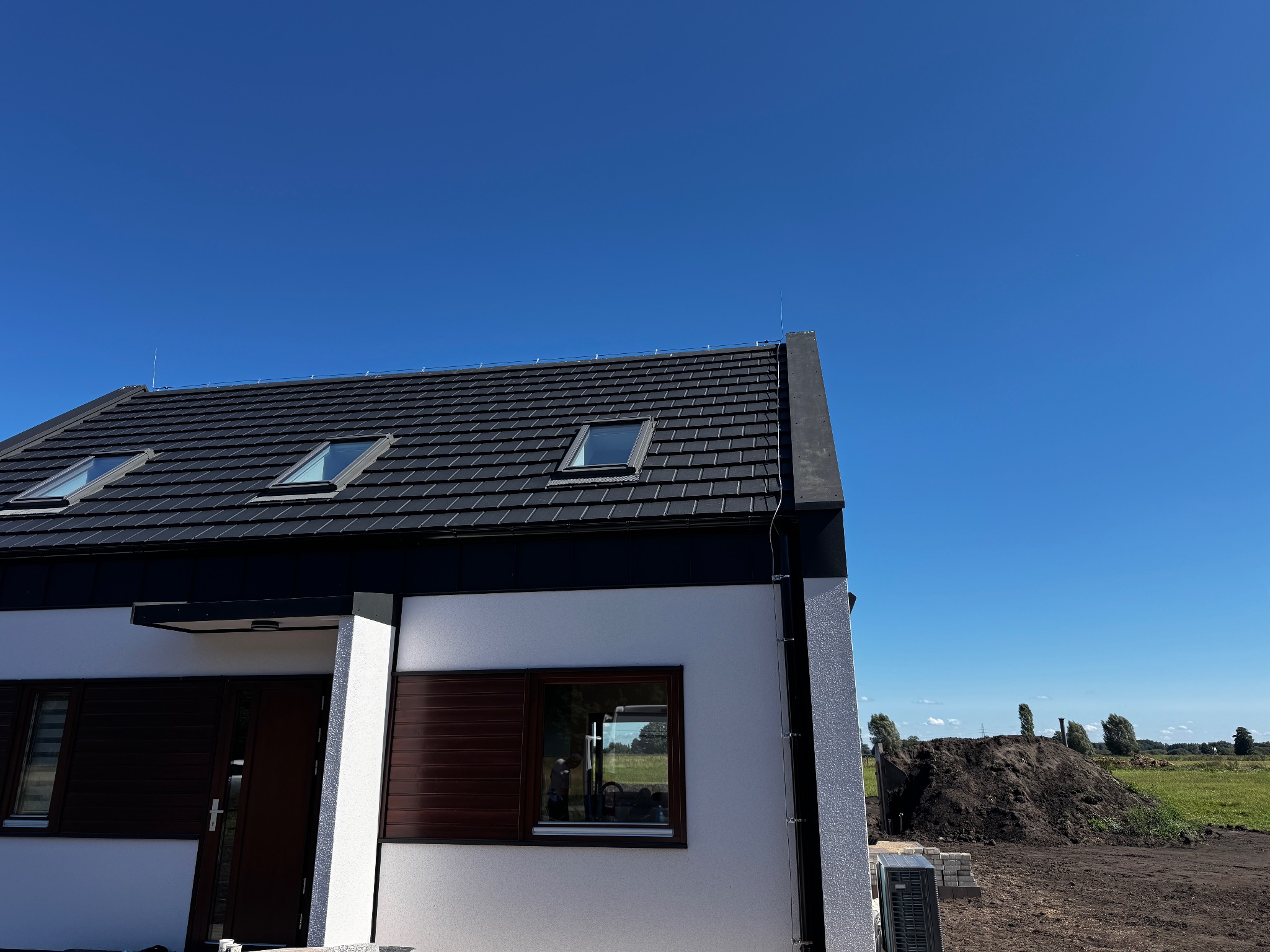 Nowoczesny dom jednorodzinny z ciemną dachówką i oknami dachowymi, na dachu widoczny maszt instalacji odgromowej, błękitne niebo w tle.