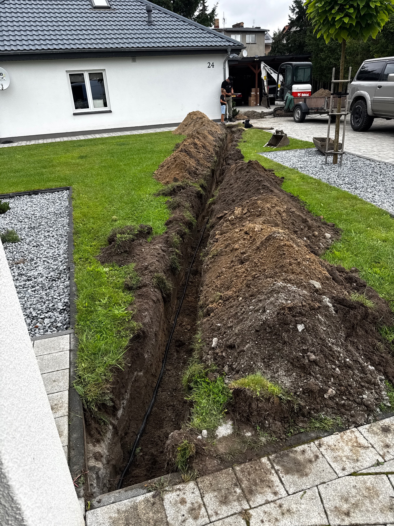 Wykop w trawniku pod kabel, widoczny czarny przewód w ziemi. W tle budynek z numerem 24, minikoparka i osoby nadzorujące prace.