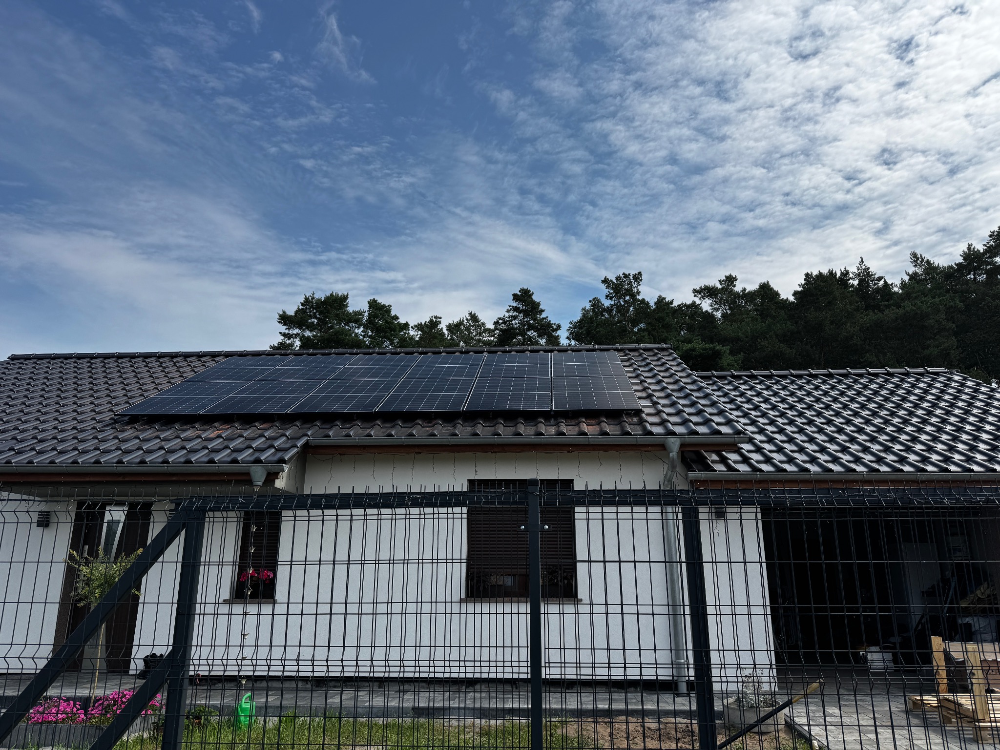 Dom jednorodzinny z panelami fotowoltaicznymi na dachu, widok zza ogrodzenia panelowego. Błękitne niebo z chmurami w tle. Ujęcie z poziomu gruntu.