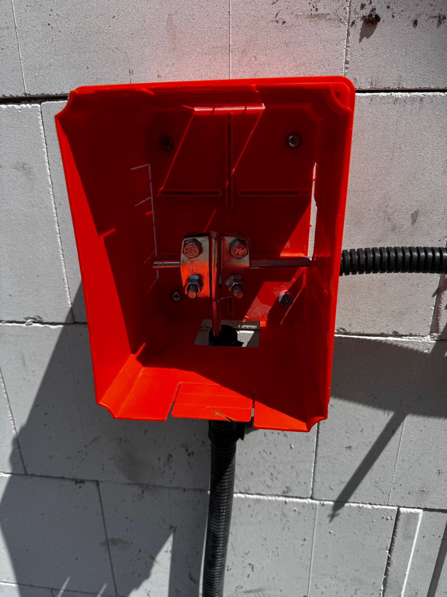 Czerwona skrzynka z metalowymi elementami i czarnym kablem na tle szarego muru z betonu komórkowego, oświetlona z lewej strony.