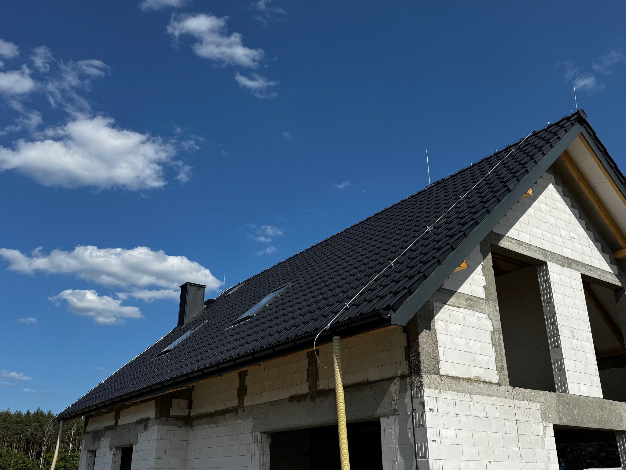 Dom w stanie surowym otwartym z czarną dachówką i zainstalowaną instalacją odgromową. Błękitne niebo z chmurami w tle.