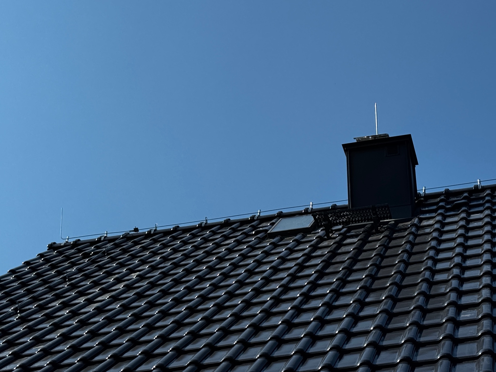 Czarny dach z dachówką ceramiczną i zainstalowaną instalacją odgromową oraz oknem dachowym, widok z dołu pod kątem na tle błękitnego nieba.