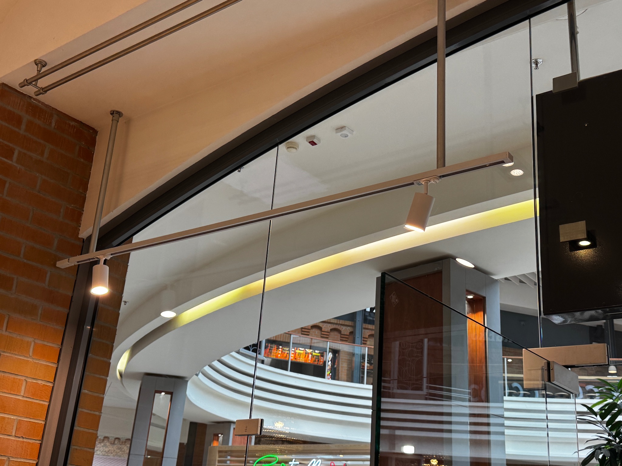 Nowoczesny system szynowy z reflektorami, zamontowany na suficie wewnątrz budynku. Widoczna instalacja oświetleniowa i fragment wnętrza z elementami cegły i szkła.