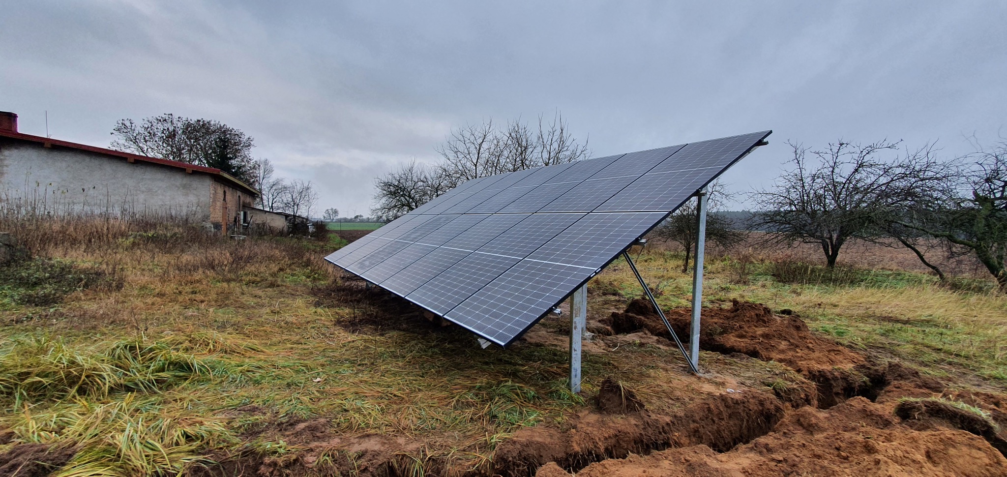 Panel fotowoltaiczny zamontowany na ziemi na metalowej konstrukcji, widoczne wykopy w ziemi, w tle budynek gospodarczy i drzewa pod pochmurnym niebem.
