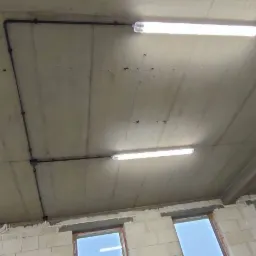 Surowy betonowy sufit z widocznymi przewodami i zamontowanymi lampami fluorescencyjnymi w nowym budynku z oknami.