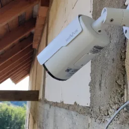 Kamera monitoringu marki Novus zamontowana na surowej ścianie budynku z widoczną konstrukcją dachu i fragmentem krajobrazu w tle.