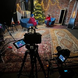 AMIX ALIAKSANDR MAROZ - Plan filmowy z dwoma osobami siedzącymi na sofach przed choinką, oświetlony lampami studyjnymi, z widocznymi kamerami na statywach i dywanem w stylu vintage.