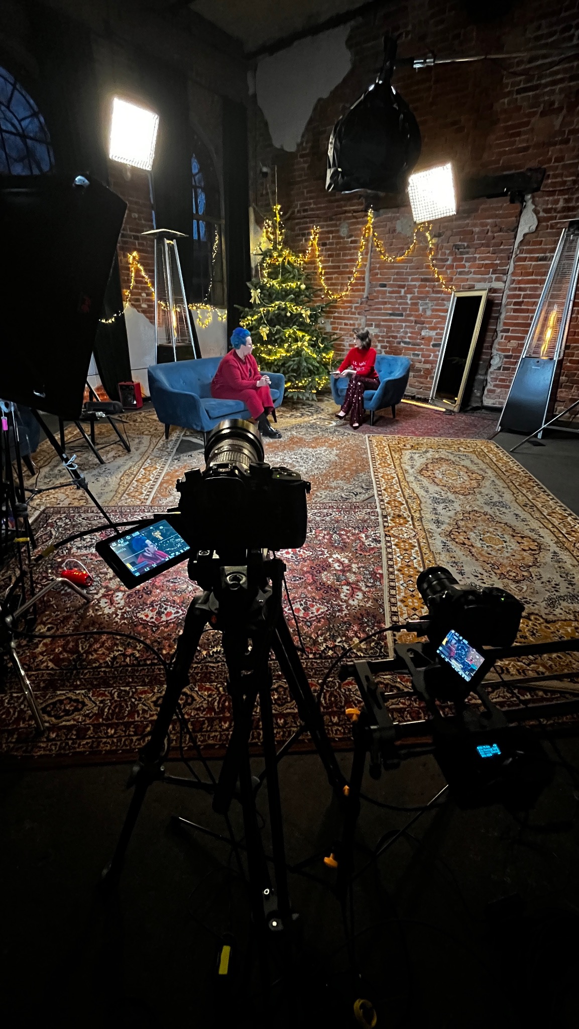 Plan filmowy z dwoma osobami siedzącymi na sofach przed choinką, oświetlony lampami studyjnymi, z widocznymi kamerami na statywach i dywanem w stylu vintage.