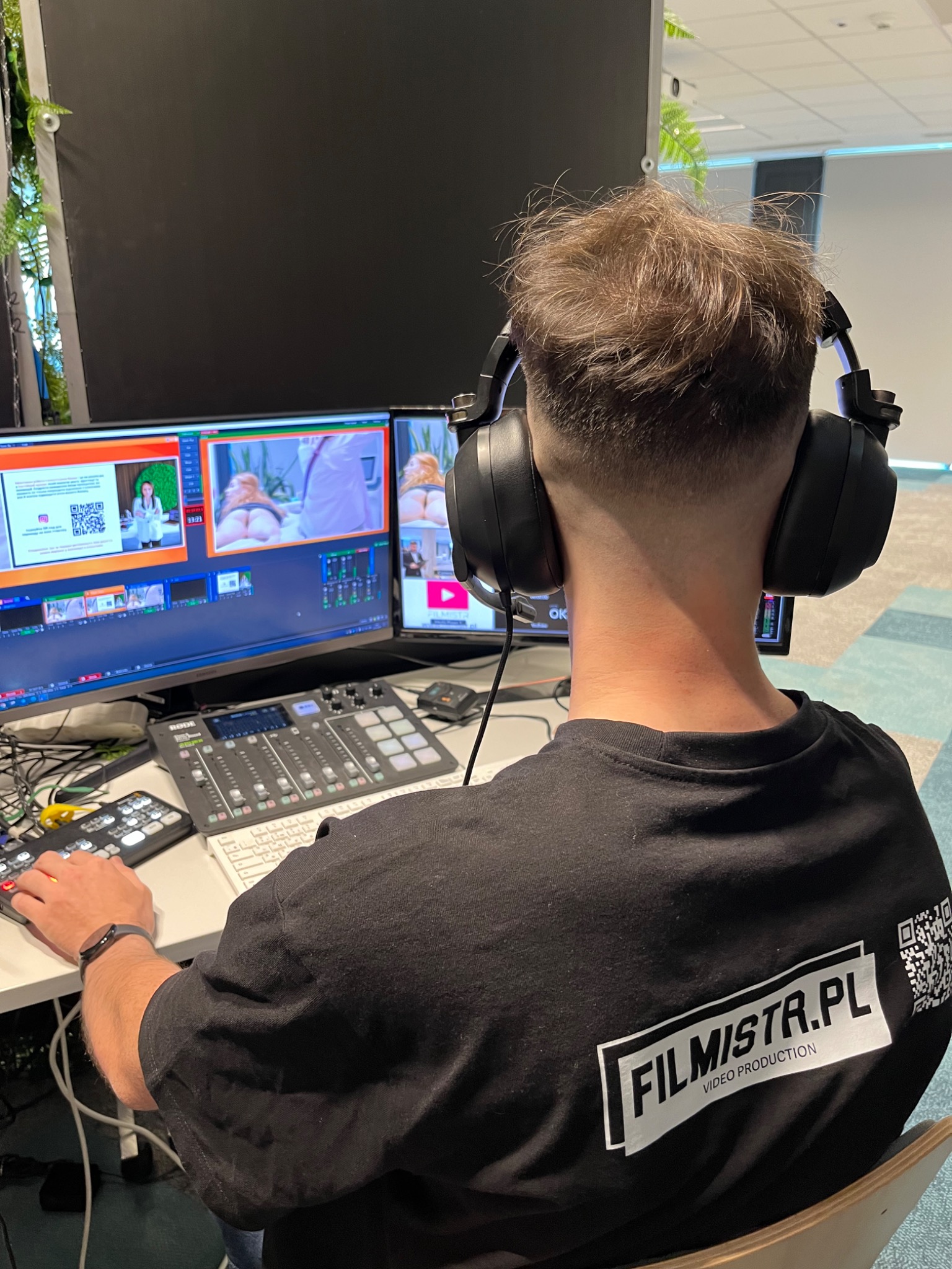 Operator w słuchawkach, plecami do kamery, edytuje materiał wideo na dwóch monitorach, z widocznym mikserem audio i logiem firmy 'FILMISTR.PL VIDEO PRODUCTION' na koszulce.
